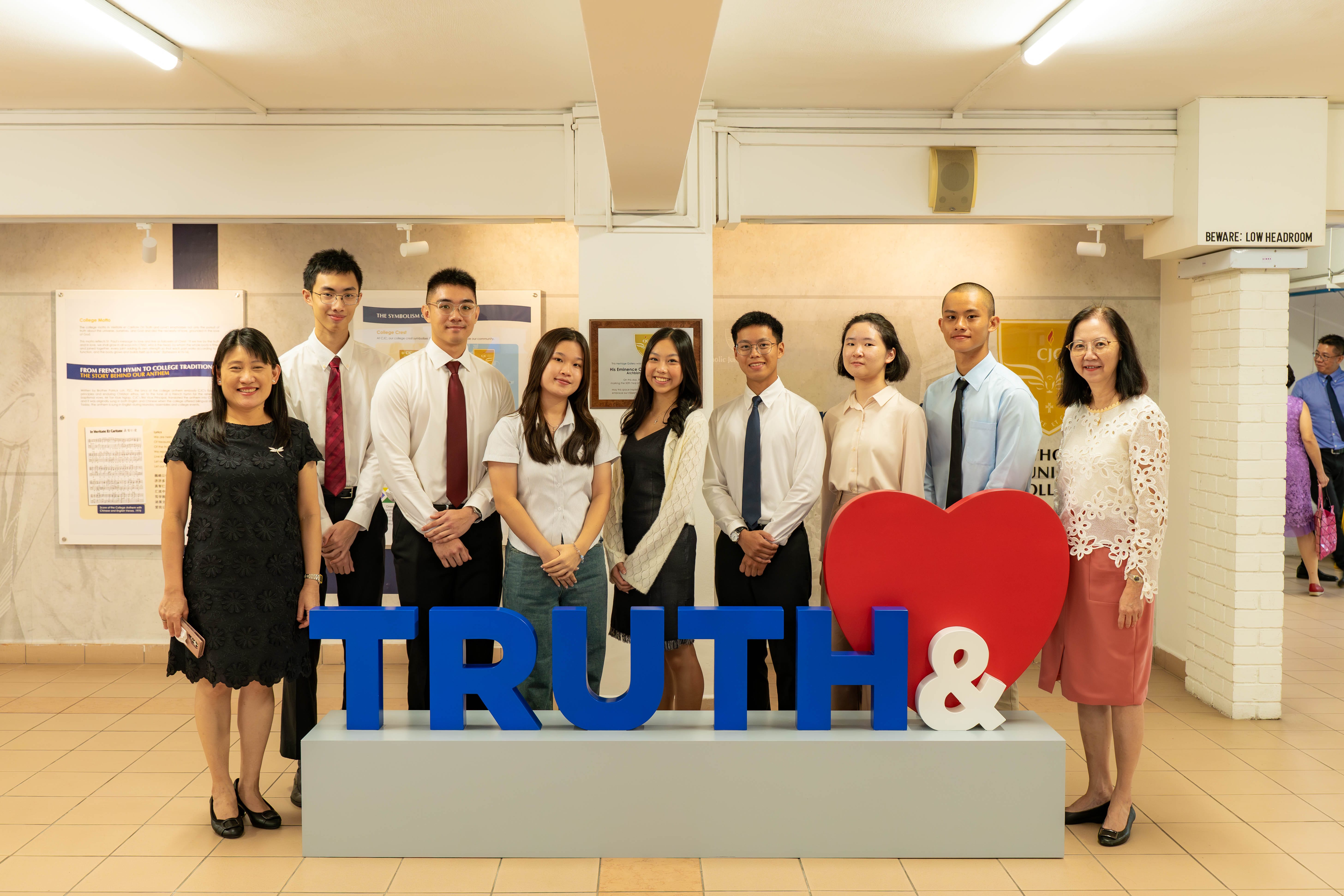 Nine people pose behind a sign reading TRUTH with a large red heart and ampersand in a college building.